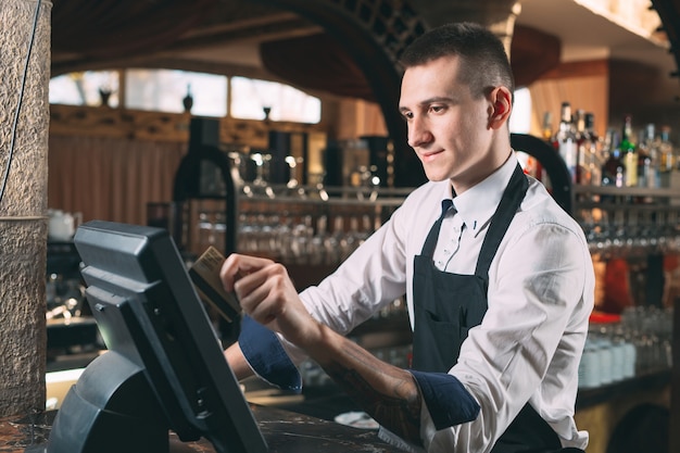 happy man or waiter in apron at counter with cashbox working at bar or coffee shop