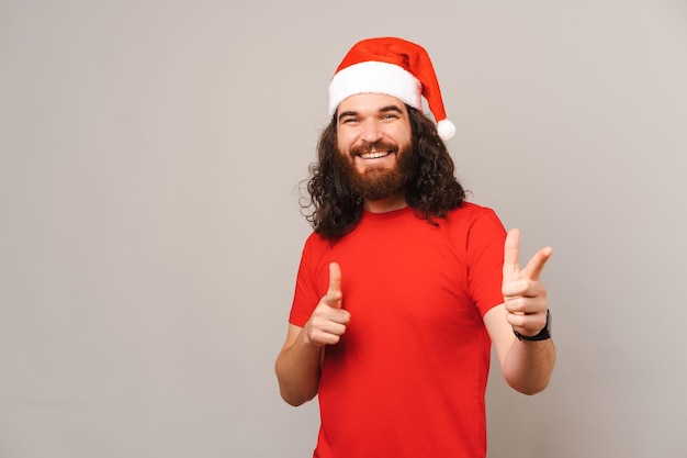 Happy man wearing Christmas hat is pointing with both hands and looking at you