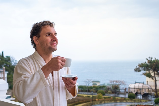 Happy man in a white bathrobe meets the morning with a cup of tea or coffee on a background of the sea. The concept of rest, health and awakening.