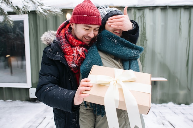 Happy man in winter clothes covering eyes of his woman, while holding the present in front of her. Standing in front of a small snowy house.