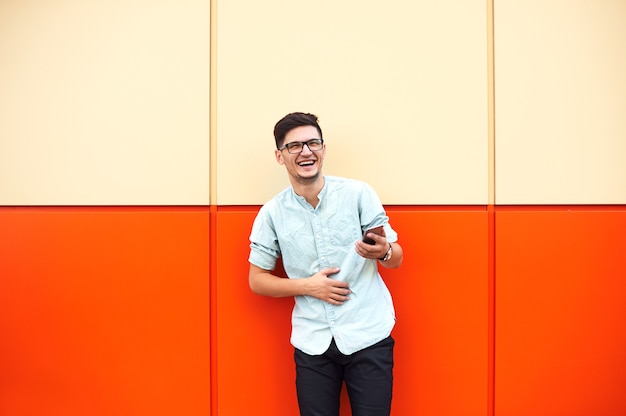 Happy man with eyeglasses smiling and using smartphone over orange wall