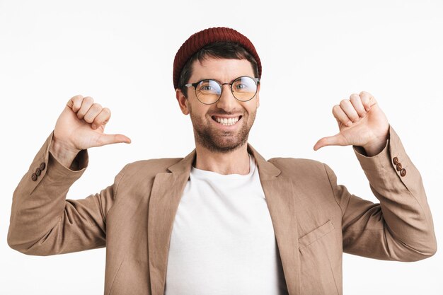 Photo happy man with stubble wearing hat and eyeglasses pointing thumbs at himself isolated over white wall