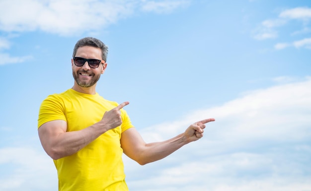 Happy man in yellow shirt and sunglasses point finger on on sky background with copy space