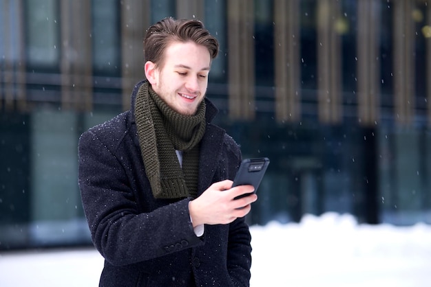 Happy man, young guy businessman is looking at screen of his cell mobile phone at winter snow day