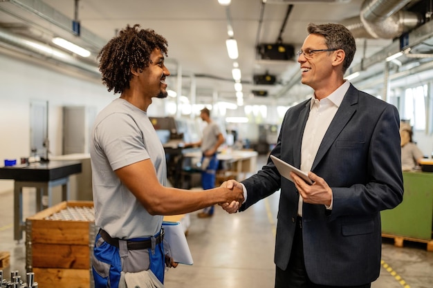 Happy manager with touchpad shaking hands with African American steel worker in a factory