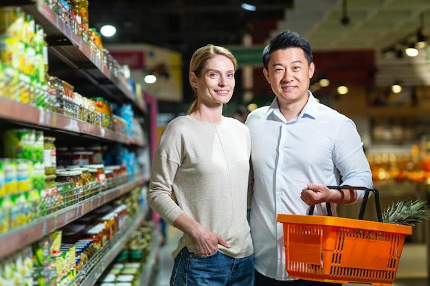 Happy married couple Asian man and woman looking at camera and smiling in grocery supermarket choosing goods