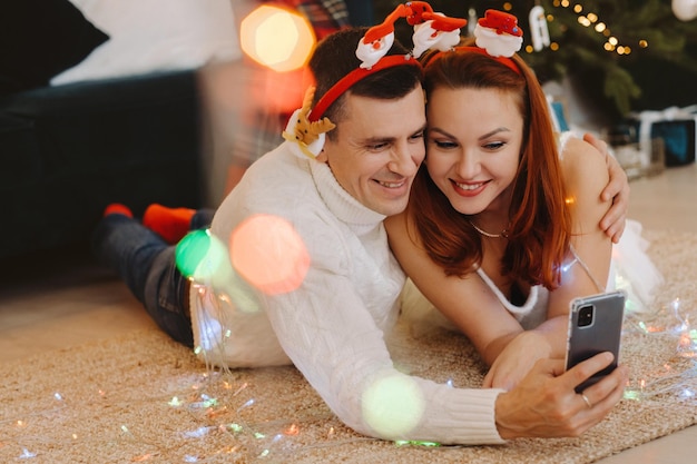 A happy married couple is lying on the floor at home near the Christmas tree and taking pictures of themselves.