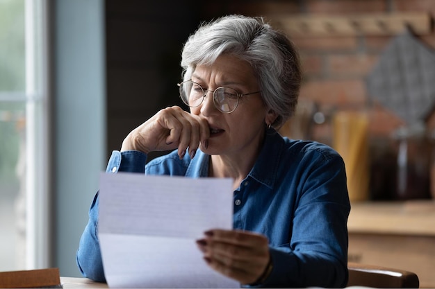 Happy mature 60s woman in glasses sit at table manage paperwork read postal letter correspondence Smiling elderly Caucasian female in spectacles consider post document or bank paper notice