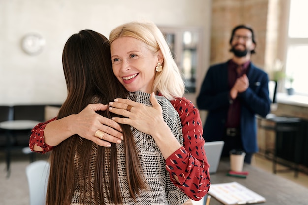 Happy mature blond businesswoman congratulating one of colleagues on great work while embracing her against young man applauding