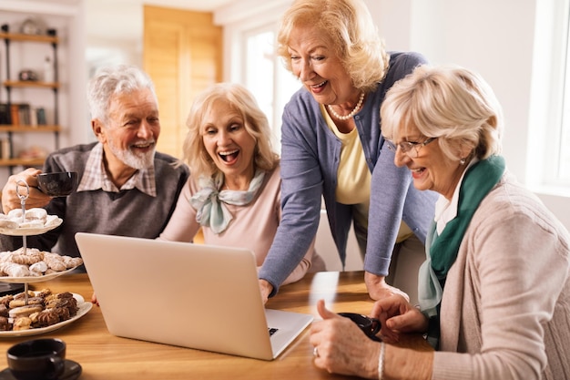 Happy mature friends using laptop together during their coffee time at home