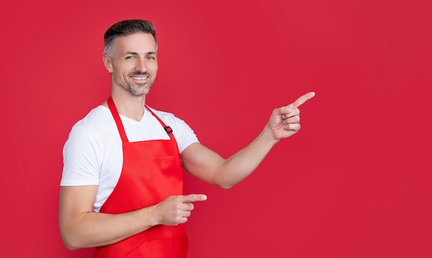 Happy mature householding man in apron on red background pointing finger