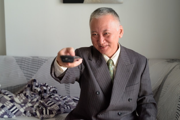 Happy mature Japanese businessman watching tv in the living room at home