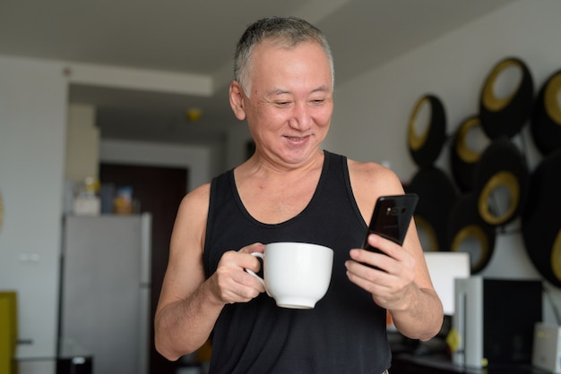 Happy mature Japanese man drinking coffee while using phone at home