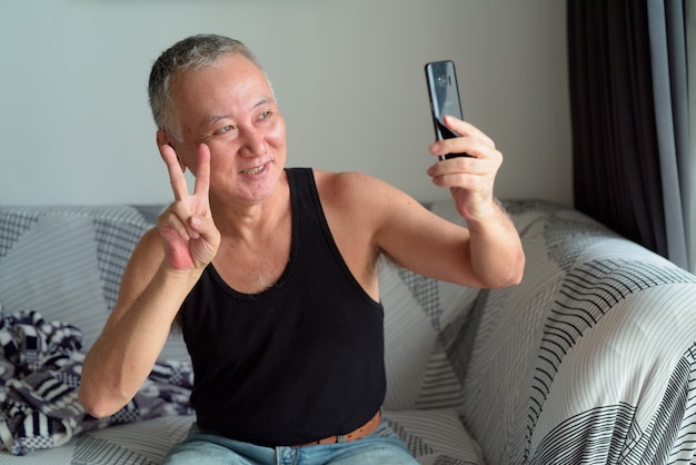 Happy mature Japanese man taking selfie in the living room at home