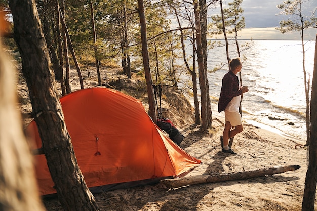 Happy mature male is spending active holiday with tent in nature and drinking coffee in morning