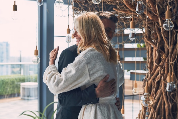 Happy mature man in full suit embracing his wife while standing indoors