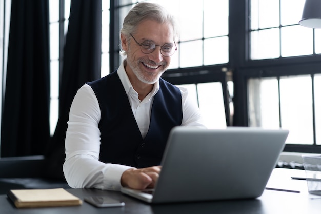 Happy mature man in full suit using laptop while working in modern office