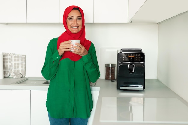 Happy mature muslim woman drinking coffee holding cup and smiling to camera standing near coffee machine in kitchen