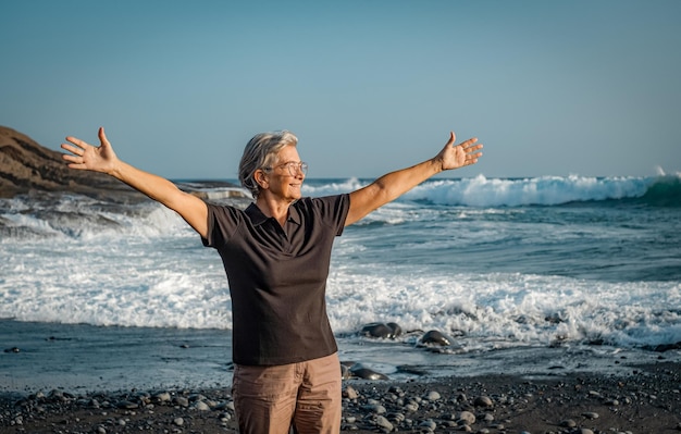 Happy mature senior woman with outstretched arms standing on the beach at sunset looking away