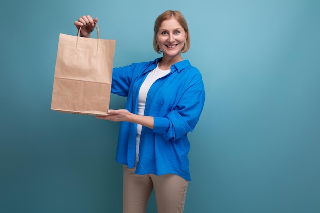 Happy mature woman holding craft bag for shopping on blue background copy space