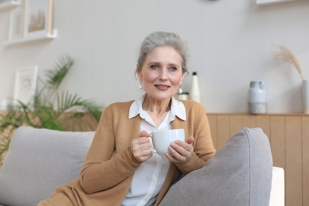 Happy mature woman resting on comfortable sofa drink coffee or tea, looking away, relaxing on cozy couch at home enjoy hot beverage