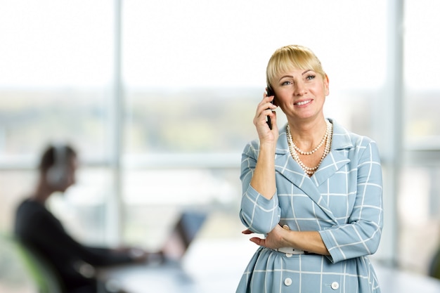 Happy mature woman talking on cell phone. Cheerful mature woman talking over phone on office window. Happy mid woman using mobile phone.