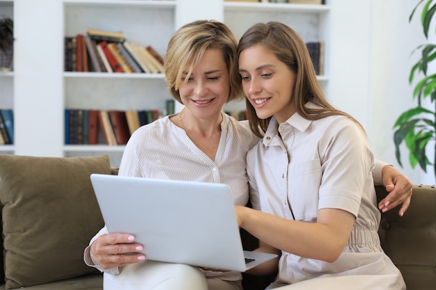 Happy mature woman with her adult daughter smiling while sitting on sofa and using laptop in living room.