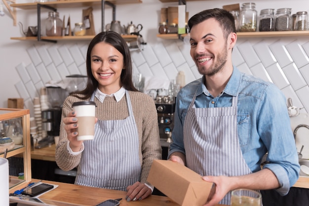 Happy meal. Pleasant young couple holding box and cup of coffee and smiling while standing behind the bar.