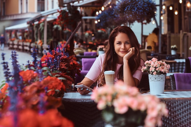 Happy middle age businesswoman with long brown hair wearing a pink dress holds smartphone while sitting at the outdoor cafe.