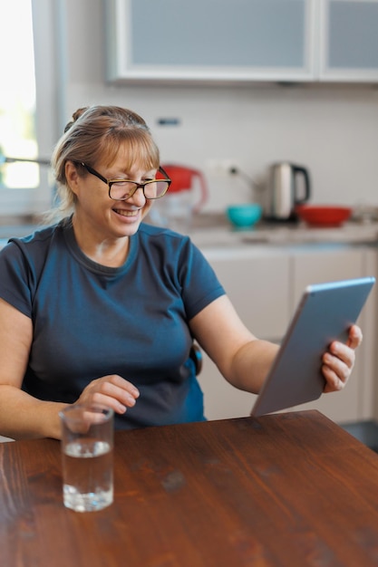 Happy middle aged 50 years old woman using digital tablet sitting in kitchen at home