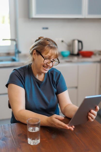 Happy middle aged 50 years old woman using digital tablet sitting in kitchen at home