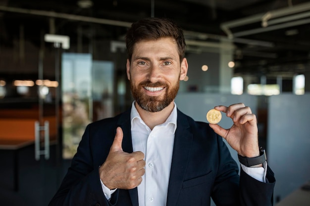 Happy middle aged businessman showing bitcoin and gesturing thumb up in modern office interior free space