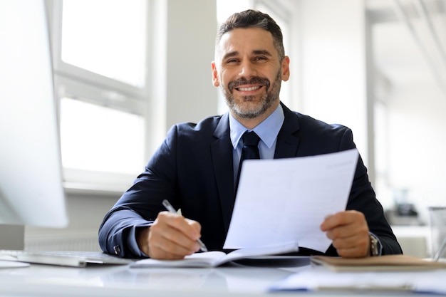 Happy middle aged businessman in suit working with documents at workplace in office and smiling at camera free space