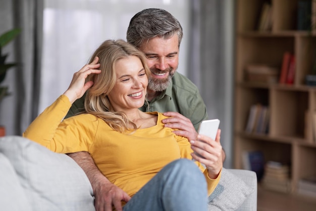 Happy middle aged couple using smartphone while relaxing on couch at home