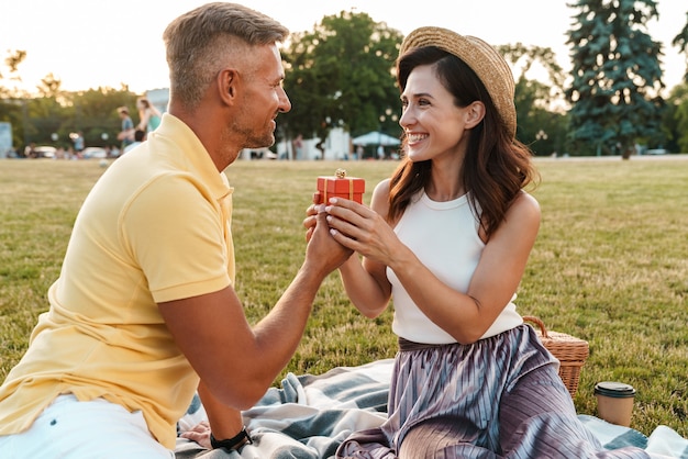 happy middle-aged man giving present box to beautiful woman while sitting on grass in summer park