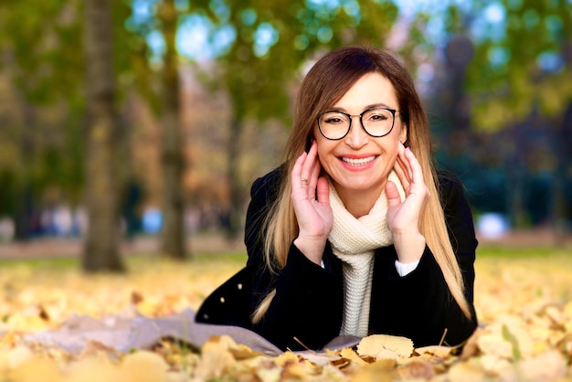 Happy middle aged mature adult woman is lying on leaves in park at golden autumn fall day, smiling