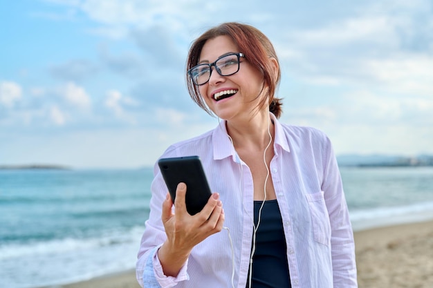 Happy middle aged woman on sea vacation enjoying in headphones with smartphone