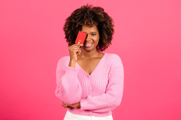 Happy millennial african american curly woman shopaholic in casual puts credit card to eye has fun