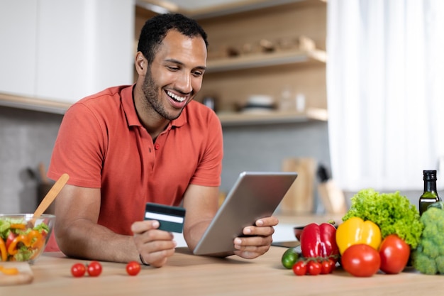 Happy millennial african american guy at table with organic vegetables uses credit card and tablet