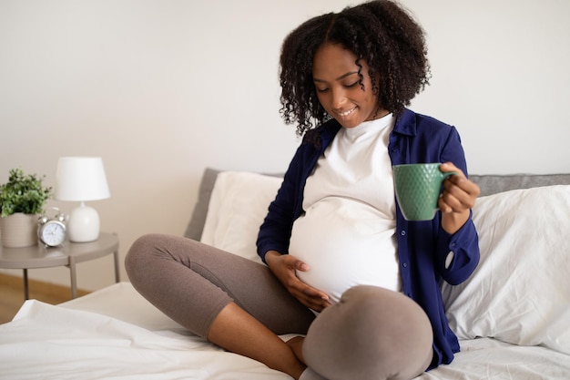 Happy millennial african american pregnant lady with big belly drinks cup of tea feels baby move on