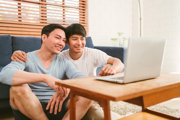 Happy millennial Asian gay couple sitting on the floor leaning on a sofa while watching content on a laptop together in the living room at home LGBT multirelationship Gay couple concept