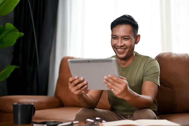 Happy millennial Asian man using his digital tablet on a sofa in the living room