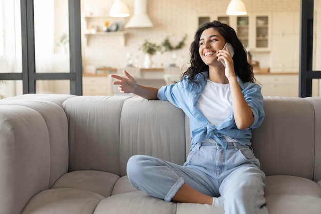 Happy millennial brunette woman sitting on sofa making call on mobile phone at home free space