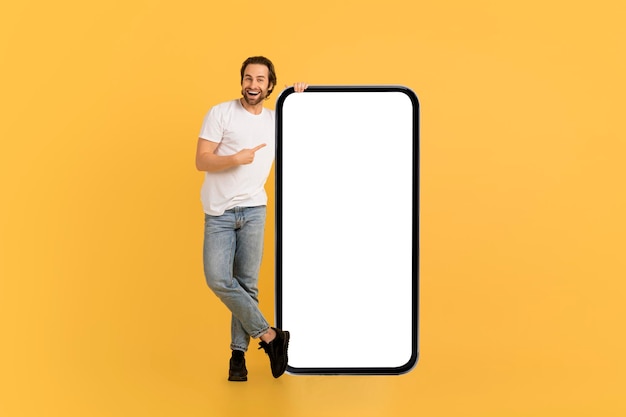 Happy millennial caucasian man in white tshirt show finger on huge smartphone with empty screen