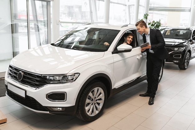 Happy millennial couple clarifying car purchase details with salesman in dealership center empty space