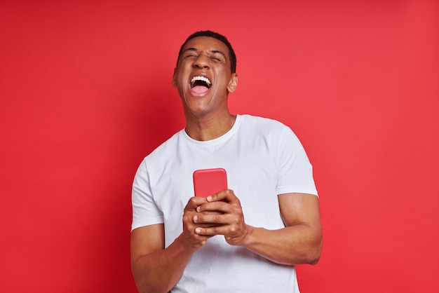 Happy mixed race man holding smart phone while standing against red background