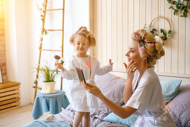 Happy mom and daughter make a make-up sitting on the bed in the bedroom