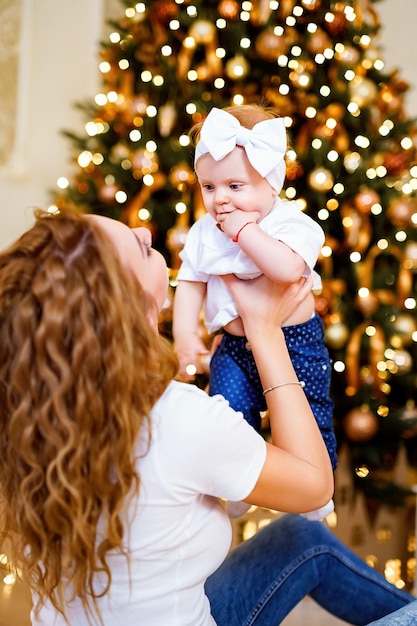 Happy mom playing with little cute baby daughter while sitting near Christmas tree in living room