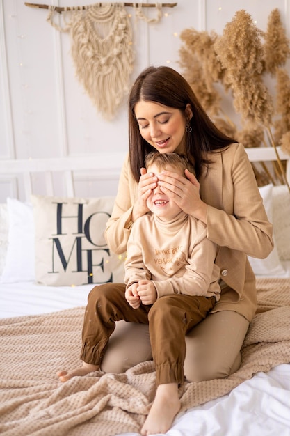 Happy mom plays peekaboo with a small child closing his eyes on a bed in beige brown natural tones having fun together