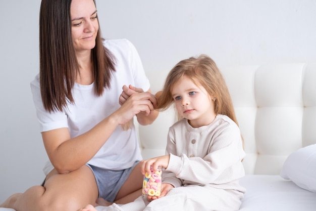 happy mother brushing daughter long hair with eco friendly wooden comb sitting on bed at home